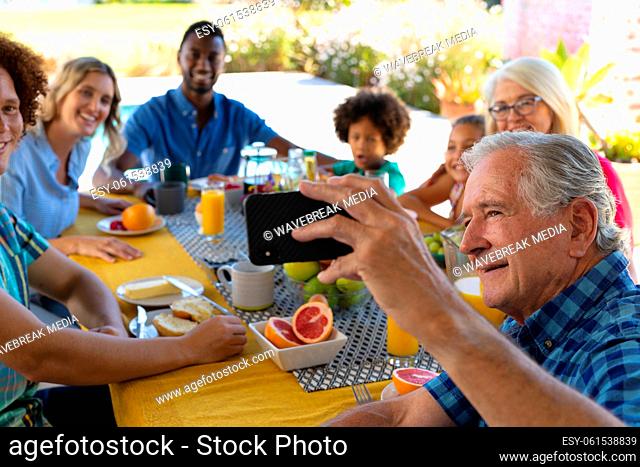 Smiling senior man taking selfie over smartphone with multiracial family while having breakfast