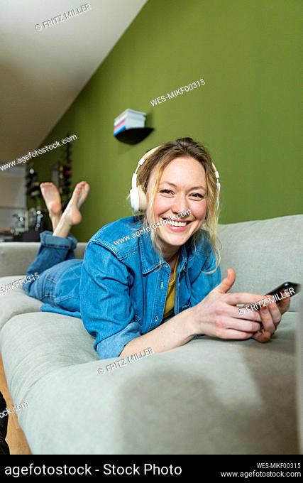 Smiling woman listening music with headphones lying on couch