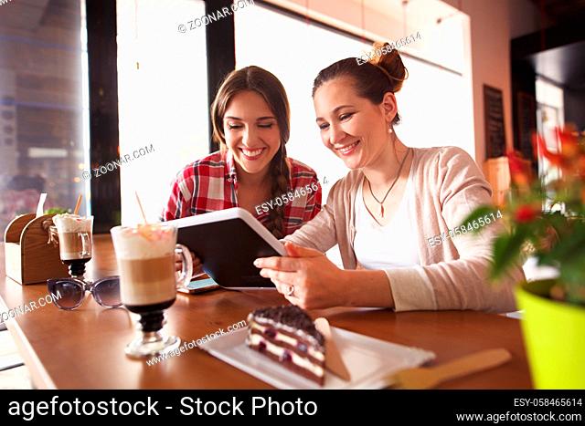Toned image of best friends girls looking at tablet PC and discussing photos or pictures. Beautiful ladies drinking coffee and eating cake