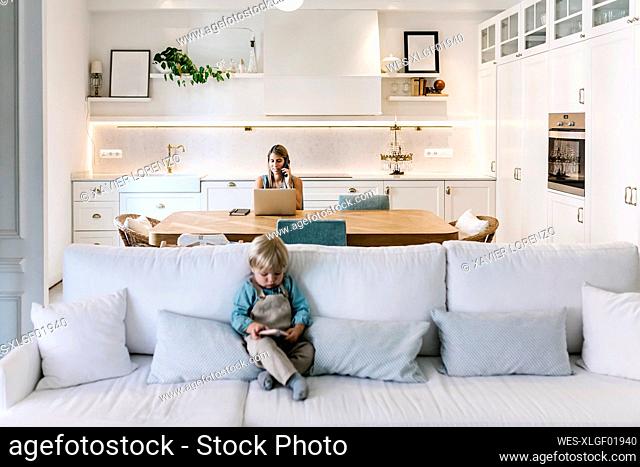 Baby boy using phone on sofa with mother working in background at kitchen