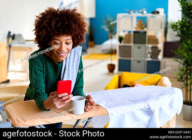 Smiling Afro woman holding coffee mug using smart phone leaning on ironing board