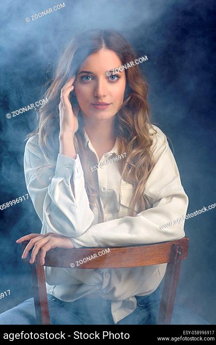 Portrait of young beautiful blonde woman is sitting on a chair. Female on smoke background