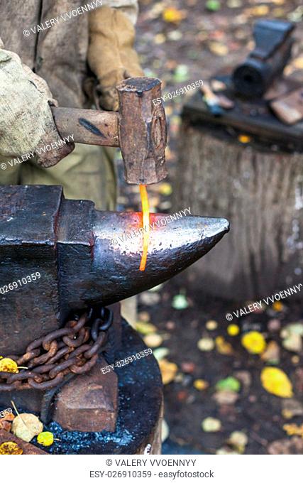 Blacksmith forges red hot glowing iron rod on anvil in outdoor rural smithy