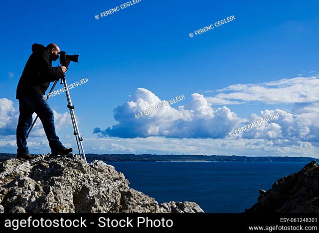 Outdoor photographer standing on a rock and taking photos of the breathtaking landscape of Brittany near Pointe de Penhir