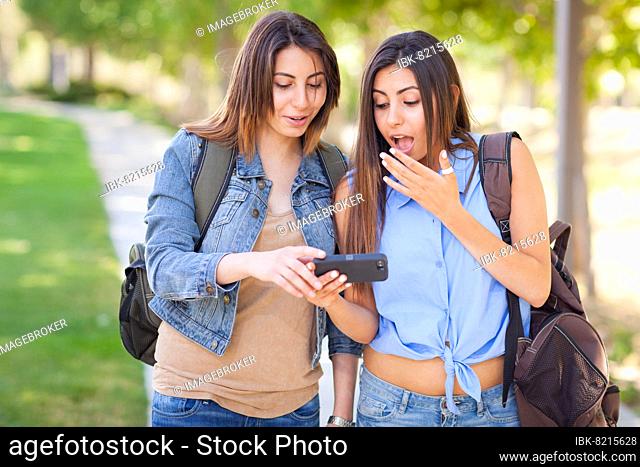 Two beautiful young ethnic twin sisters with backpacks using A smartphone outside