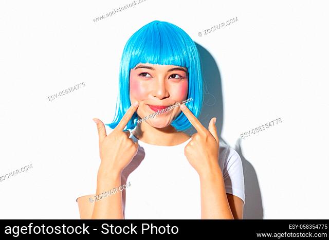 Image of adorable asian girl in blue wig poking her cheeks and looking upper left corner, standing over white background
