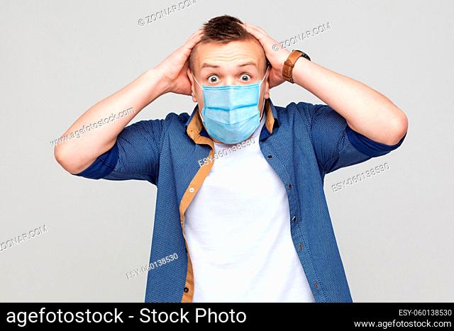 Oh my God. Portrait of shocked young man in casual style with surgical medical mask standing, holding head and looking at camera with unbelievable face
