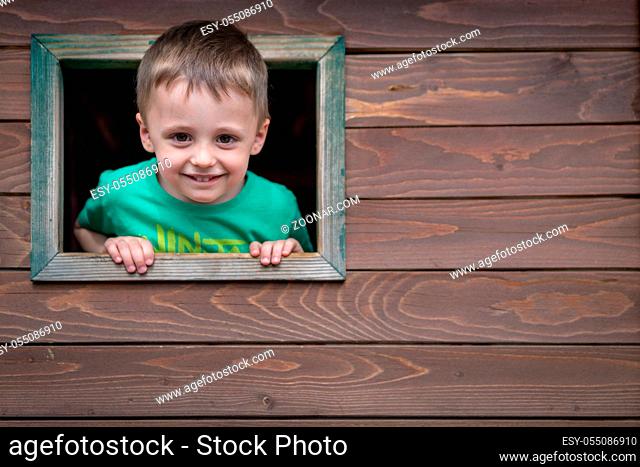 Portrait of a cute little Caucasian boy looking through the window of a wooden toy house in a outdoor playground