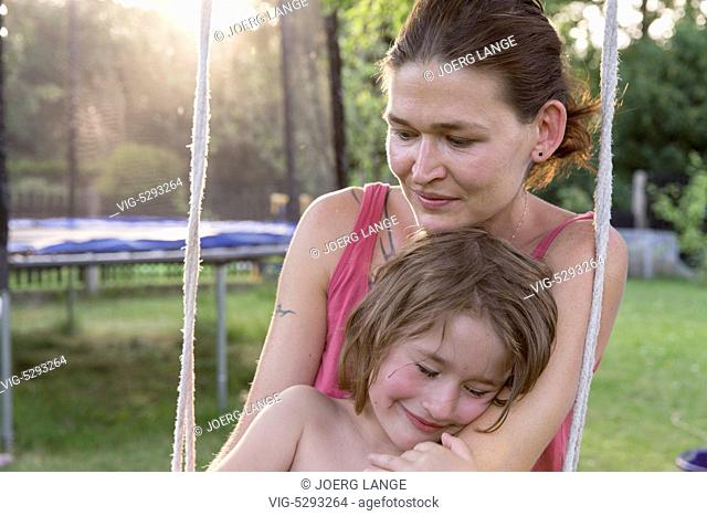Mother sits in the sunshine on a swing in the garden and holds her daugther lovingly.- - Germany, 21/09/2010