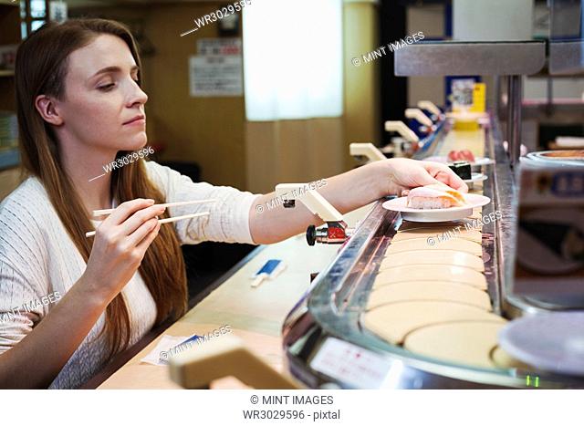 Young Caucasian woman sitting in a sushi bar, choosing a dish from a sushi train, Kaiten-zushi