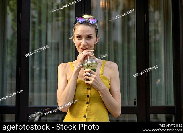 Young woman drinking smoothie while standing in front of door