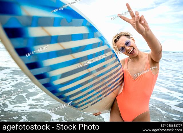 Joyful blonde girl with short hairstyle stands on the beach on the background of the sea and cloudy sky. She wears orange swimsuit with sunglasses and holds a...
