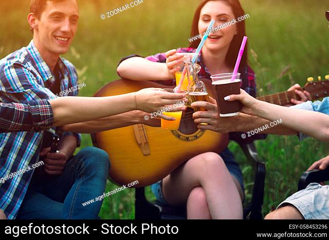 Three people toasting their beverages in park. Girl with guitar sitting in grass field next to two of her friends cheering drinks