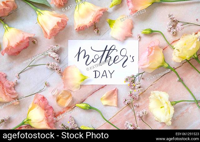 Card with the handwritten text MOTHER'S DAY surrounded by pink flowers, earrings, petals and parfume flacon top view on a marble table