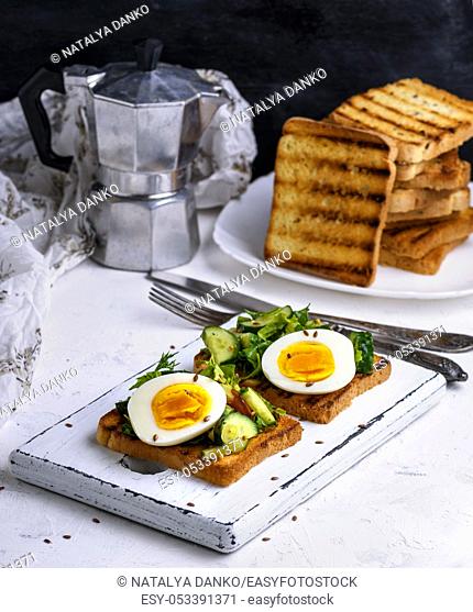 toasted square pieces of bread from white wheat flour with boiled egg, cucumber and green spinach leaves on a white wooden board, breakfast sandwich