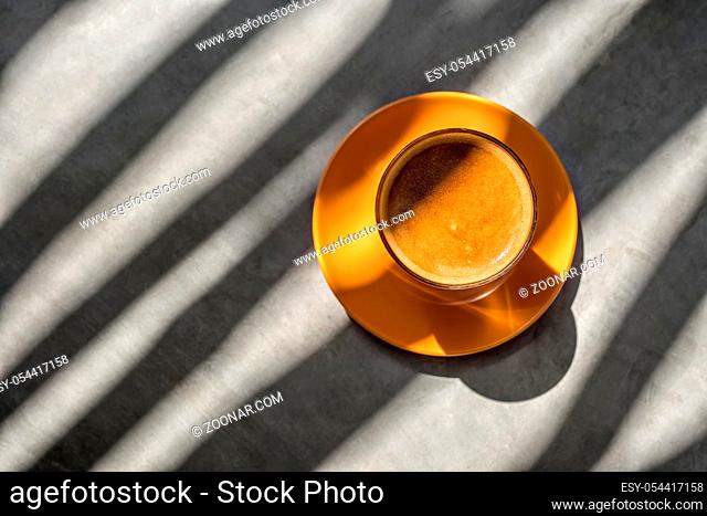 Delicious latte drink in the glass on the orange saucer on the table. Sun shines onto them and creates glares and shadow patterns. Closeup