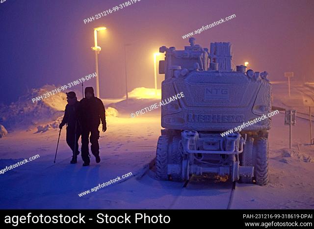 16 December 2023, Saxony-Anhalt, Schierke: Hikers walk along an excavator covered in hoarfrost on the Brocken in the early hours of the morning