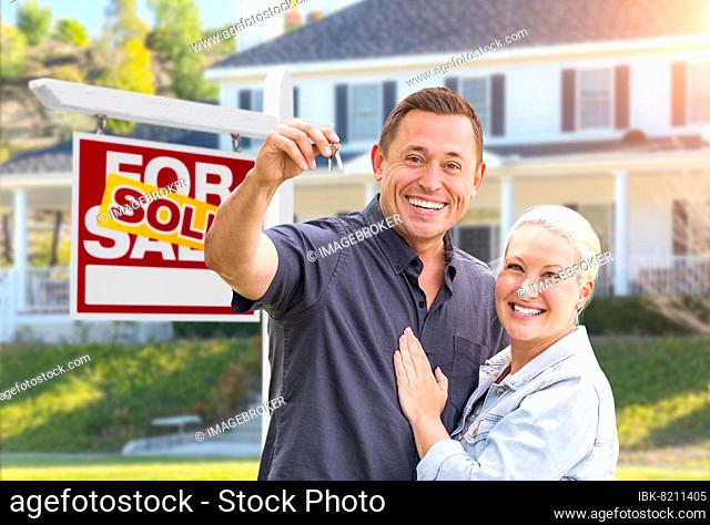 Happy couple with new house keys in front of sold real estate sign and beautiful house