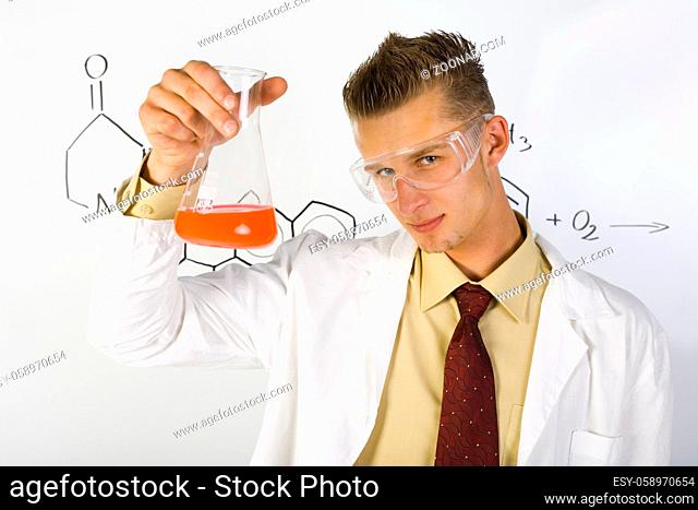 Young chemist in white apron. Holding beaker with orange liquid. Wearing goggles. Looking at camera, gray background