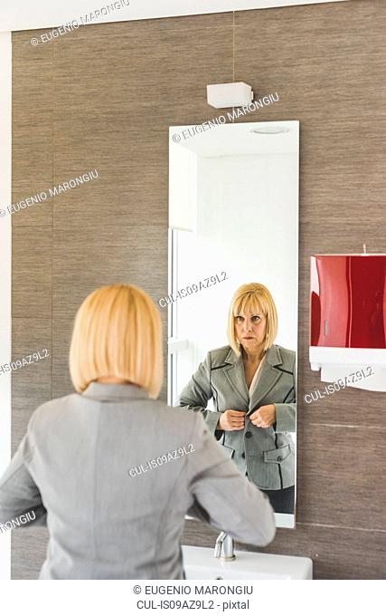 Mature businesswoman fastening jacket whilst looking in office powder room mirror