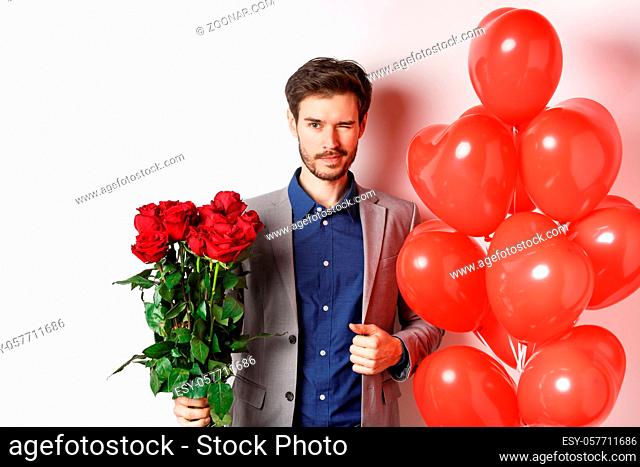 Confident boyfriend in suit going on romantic date, winking at camera, holding bouquet of red roses, standing near heart balloon and winking
