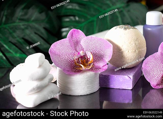 Beautiful lilac orchid flower, cosmetic bottle, cotton pads and handmade soap with stack of white stones and monstera leaves on black surface