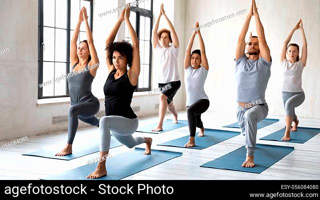 Diverse young sporty people doing Warrior one exercise at group lesson, practicing yoga in modern fitness center, standing in Virabhadrasana pose