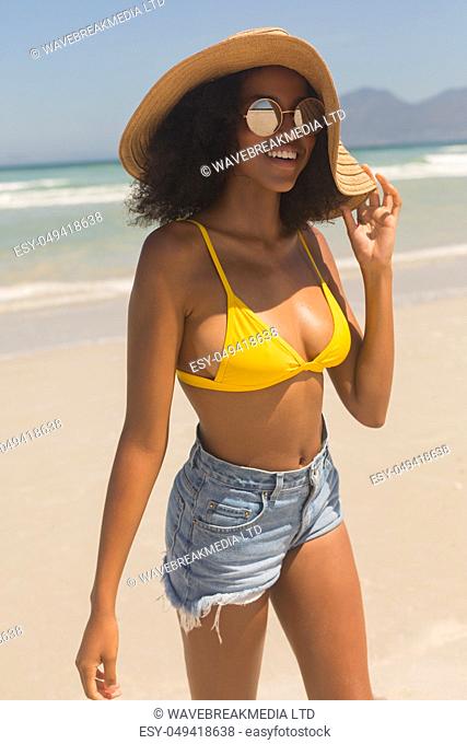 Portrait of happy young African American woman in yellow bikini, hat and sunglasses standing on the beach. She is smiling
