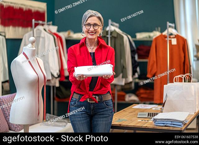 Good moments. Smiling gray-haired woman in glasses in stylish clothes pointing at camera with bundle standing in her sewing workshop
