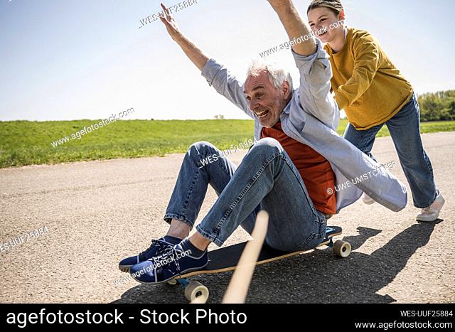 Playful girl pushing cheerful grandfather sitting with arms raised on skateboard