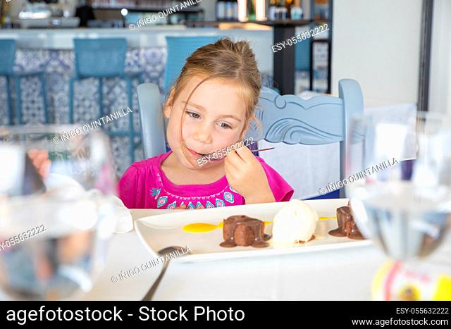 four years old blonde girl eating sweet dessert: black chocolate, vanilla ice cream, strawberries and mango sauce. Satisfaction face expression with spoon in...