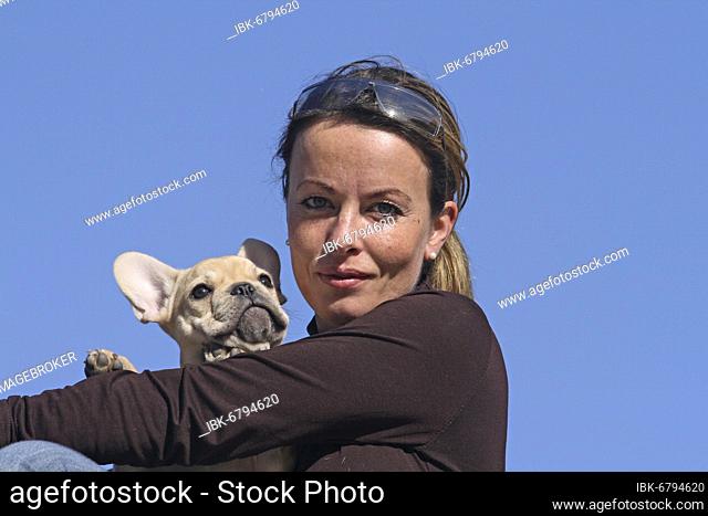Young blond woman holding 12-week-old French bulldog in her arms. Blue sky in the background Further motifs on this theme are available on request