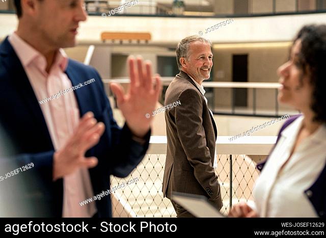 Smiling businessman standing by railing with colleagues discussing in foreground at office