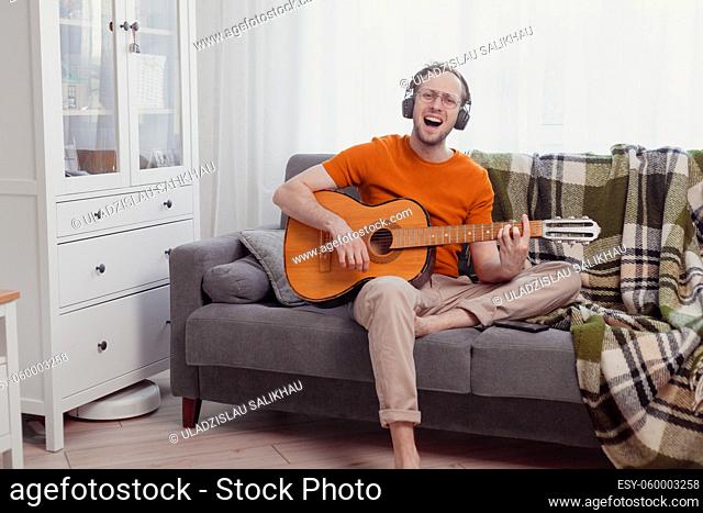 Young caucasian man in headphones playing the guitar and singing at home. Quarantine and hobbies concept. Practice music, lifestyle photo