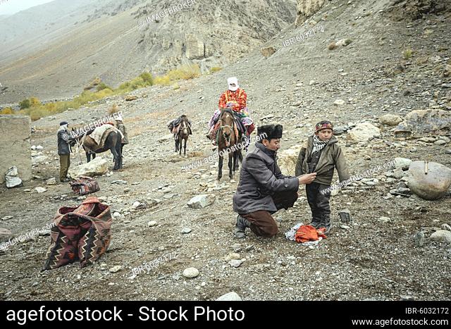 A man puts on a jacket for his son, a horse is loaded in the background, a woman sits on a horse, Kyrgyz nomads on their way to the hospital of Ishkashim