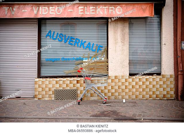 An empty shopping cart on the sidewalk in front of a closed electric goods shop with broken window pane