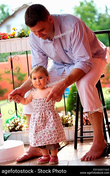 Father supports his baby girl in her first steps at home, outdoor in the garden