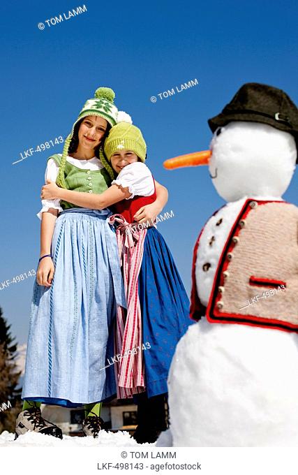 Zwei girls wearing dirndls embracing each other, snowman in foreground, Frauenalpe, Murau, Styria, Austria