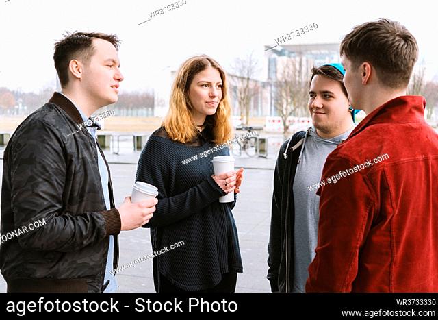 group of teenage friends having a conversation while standing together on city street holding coffee cups