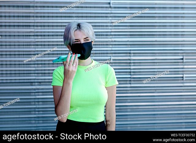 Blond woman standing in front of metal background, wearing face mask, using smartphone
