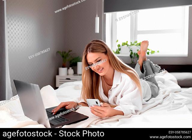 Pretty smiling Caucasian girl lays on bed in bedroom, use notebook and smartphone for online shopping. Online shopping, communication, technology