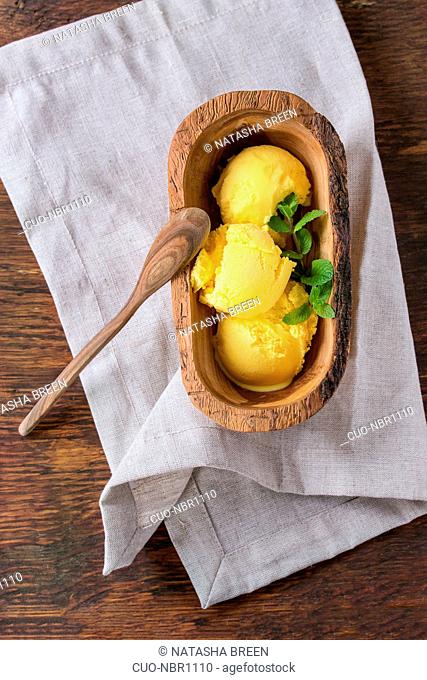 Homemade mango ice cream with fresh mint in olive wood bowl with wooden spoon, served on white textile napkin over wooden textured background