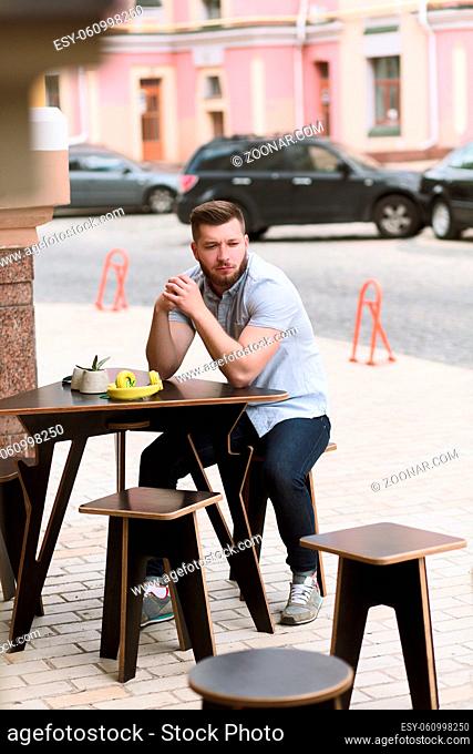 Youg man waiting for his order or friends. Handsome man sitting at wooden table in outdoor vegan cafe and looking at camera