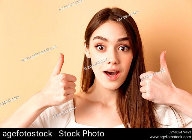 Very cool. Excited girl showing thumbs up and gasping amazed, praising good job, compliment something awesome, standing on beige background