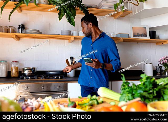 Happy african american man cooking dinner in kitchen, using smartphone