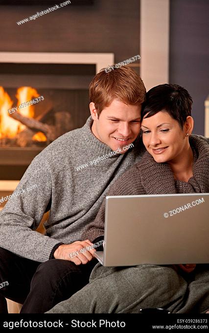 Young couple hugging on sofa at home in winter, using laptop computer, smiling