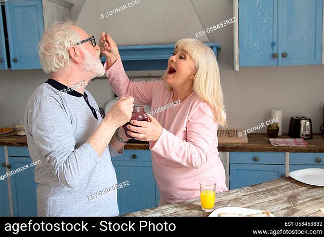 Senior couple trying out jam during breakfast. Cute old lady acting babyish giving her husband spoonful of jam in their modern designed kitchen in morning