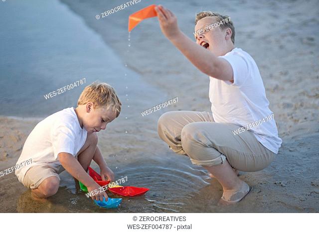 Two boys playing with paper boats at a water pool on a sandy beach