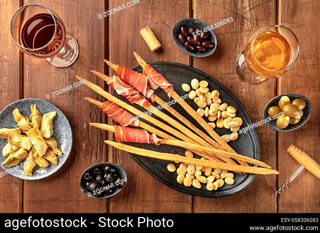 Italian antipasti. Grissini breadsticks with parma ham and roasted almonds, with olives and artichokes, shot from the top on a dark rustic wooden background...