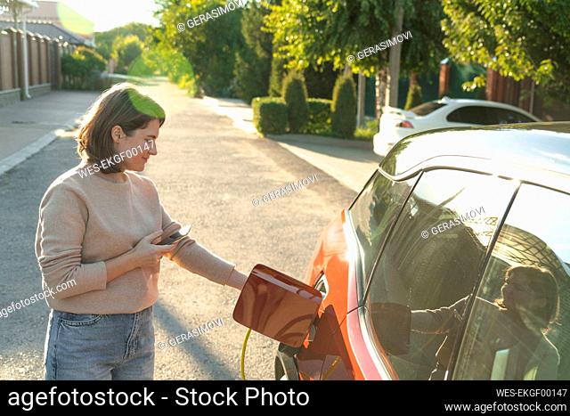 Woman with smart phone charging car at electric charging station